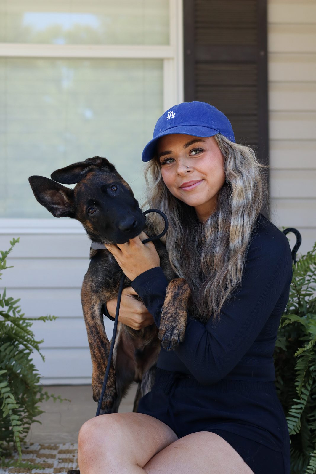 Woman in a blue cap holds a brindle Malinois puppy on a porch next to ferns