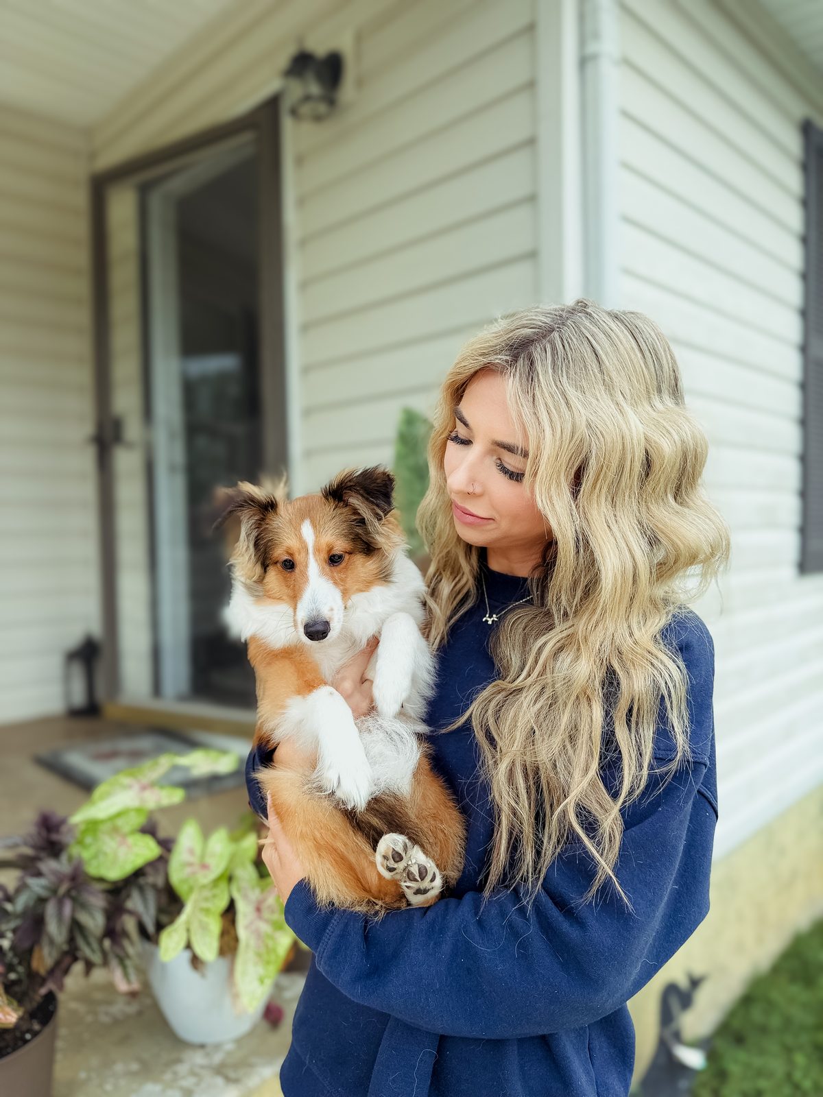 A woman holding a sheltie puppy on a front porch beside leafy plants